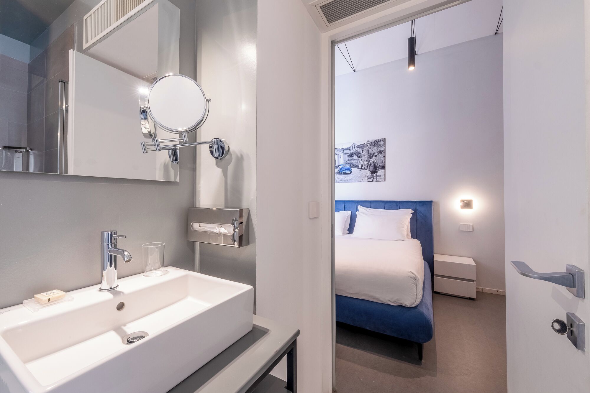 A modern bathroom with a white sink, a mirror, and a shower. The bathroom leads to a bedroom with a bed and a nightstand.