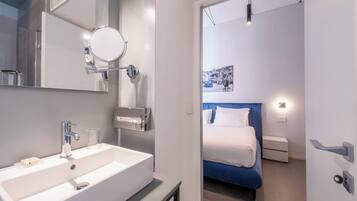 A modern bathroom with a white sink, a mirror, and a shower. The bathroom leads to a bedroom with a bed and a nightstand.