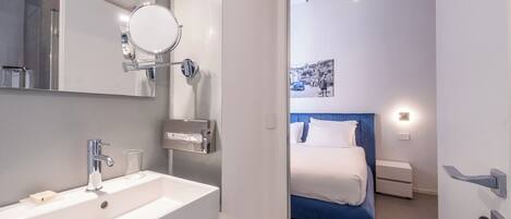 A modern bathroom with a white sink, a mirror, and a shower. The bathroom leads to a bedroom with a bed and a nightstand.