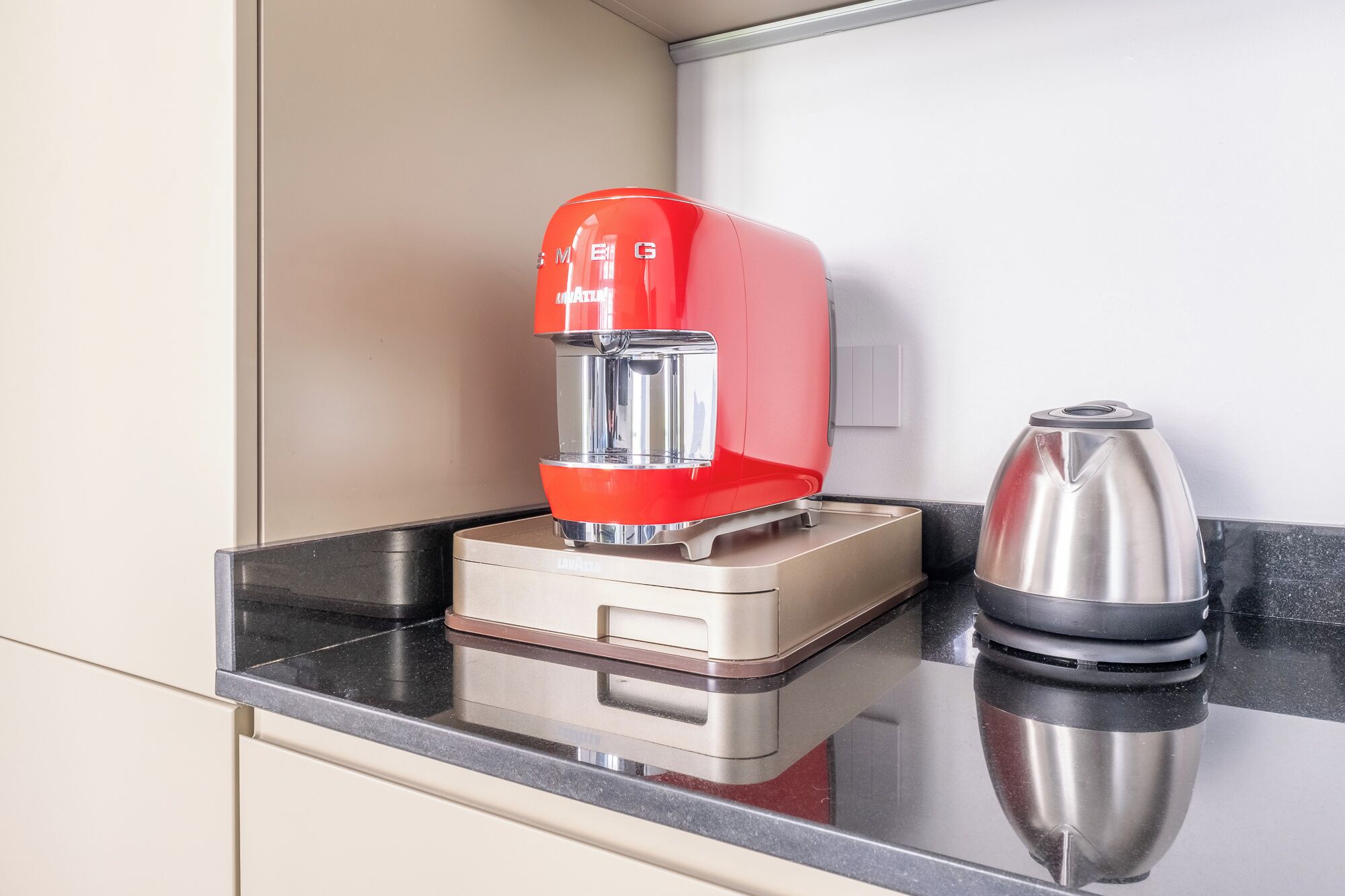 A kitchen with a red coffee maker, a stainless steel kettle, and a beige countertop.