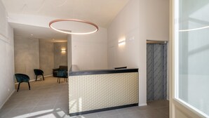 A modern reception area with a light-colored reception desk, a circular pendant light, and a view of a hallway with chairs and a door. - Oriana Homèl Torino (Turin)