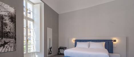Une chambre à coucher avec un grand lit, des tables de chevet, un banc, un miroir et une fenêtre donnant sur des immeubles.