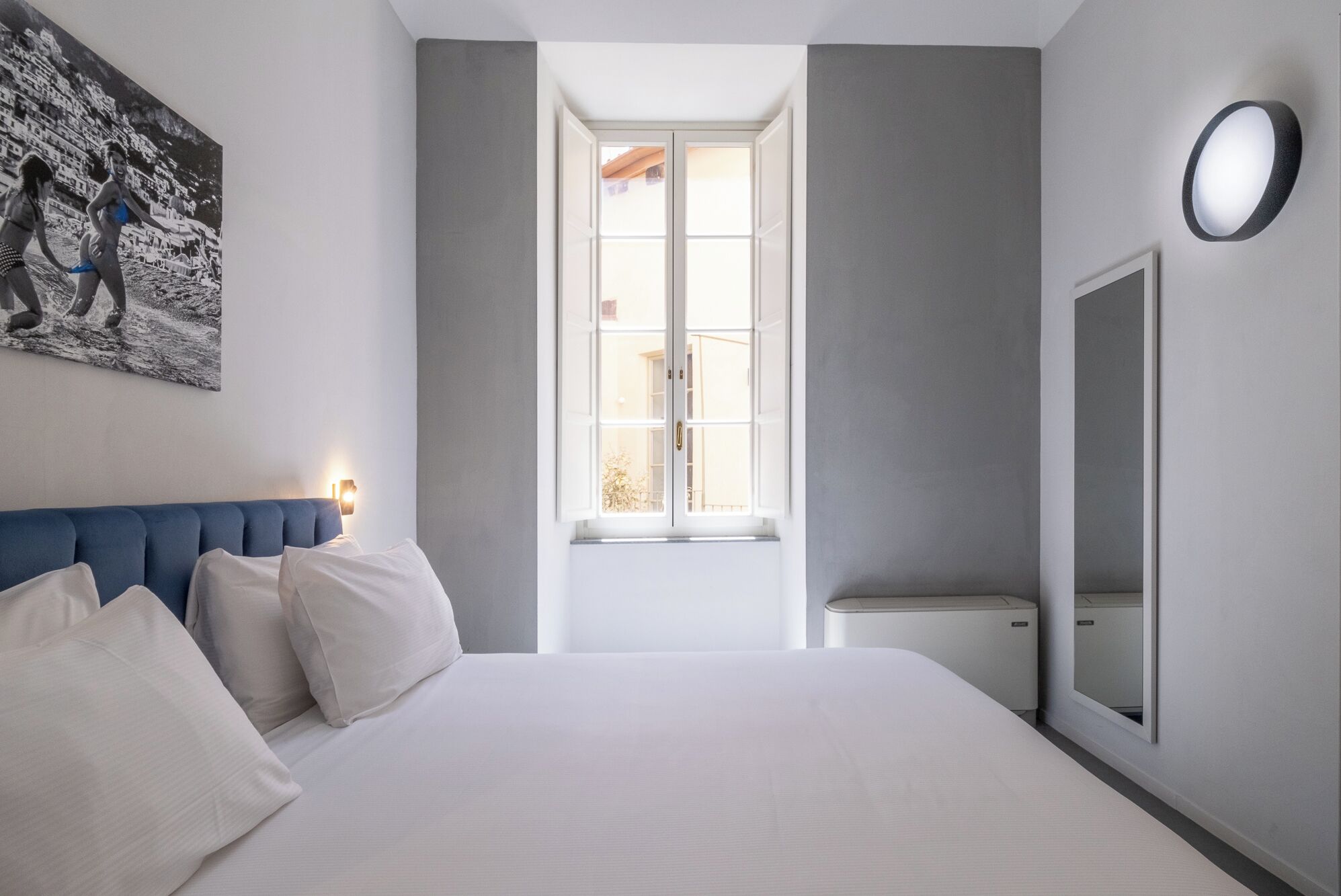 A bedroom with a large bed, a window, a wall-mounted mirror, and a painting on the wall.