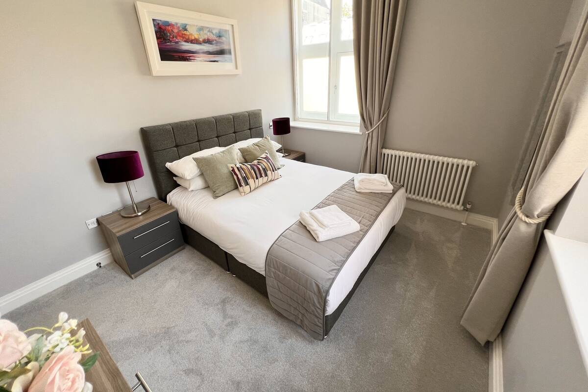 A neatly arranged bedroom with a bed, bedside tables, a lamp, a radiator, and a window with curtains.