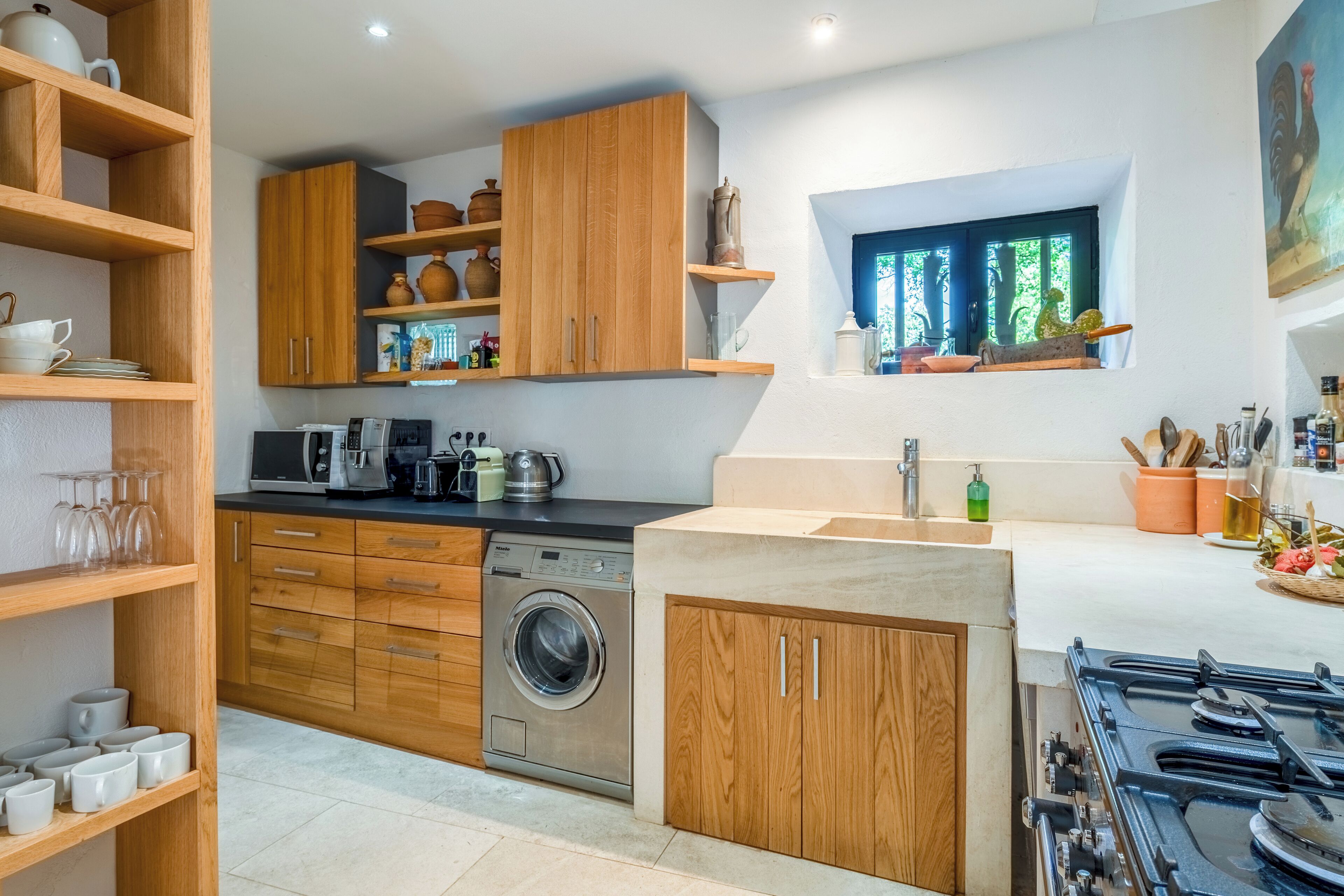A compact kitchen with wooden cabinets, a washing machine, a stove, and a window with a view.