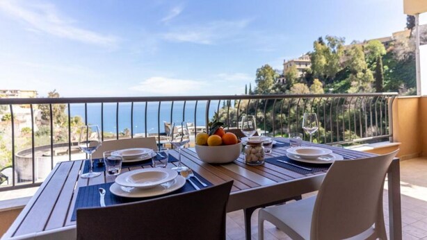 A dining table set with plates, glasses, and a fruit bowl on a balcony with a view of the sea and buildings.