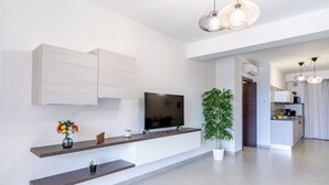 A modern living room with a flat-screen TV, built-in shelves, a potted plant, and a kitchen area in the background.