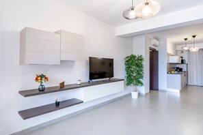 Un salon moderne avec un téléviseur à écran plat, des étagères intégrées, une plante en pot et un coin cuisine à l’arrière-plan.