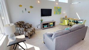 Una sala moderna con un sofá gris, un sillón estampado y un televisor de pantalla plana montado en la pared. Hay una zona de comedor con sillas amarillas y una mesa, además de decoración en la pared.