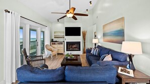A living room with a blue sofa, a fireplace, a ceiling fan, and a view of the ocean through a sliding door. - Gulffront w/ Screened Porch | Dogs Allowed (Indian Pass)