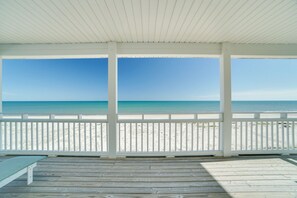 Une terrasse en bois couverte, offrant une vue sur une plage de sable et un océan d’un bleu limpide.