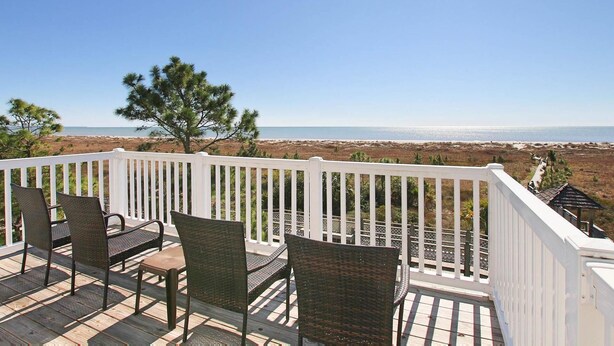A deck with wicker chairs and a table, overlooking a beach and ocean.