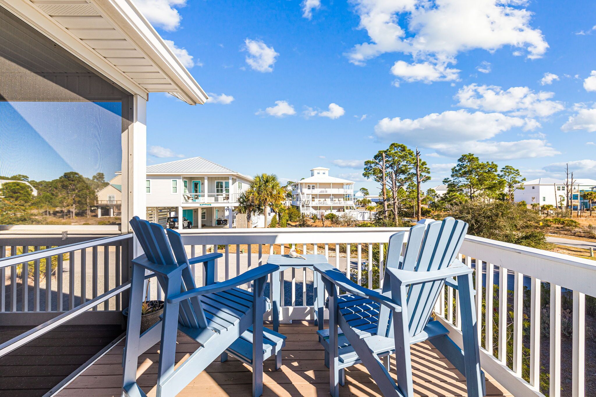 Een houten terras met blauwe Adirondack-stoelen en een witte reling, met uitzicht op een strand met huizen en palmbomen.