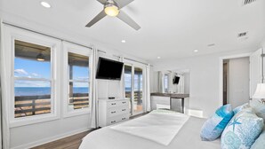 Un dormitorio con una cama grande, televisión, ventilador de techo y vistas al océano a través de las ventanas.
