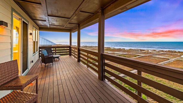 Terraza de madera con mobiliario de mimbre que da a una playa bajo un cielo con una puesta de sol colorida.