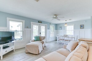 Un salon spacieux doté d’un téléviseur, de canapés beiges, d’un coin repas avec des meubles blancs et de grandes fenêtres laissant entrer la lumière naturelle.