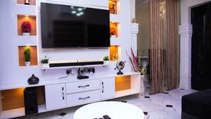 A modern living room with a large flat-screen TV mounted on a white wall, a white coffee table with remote controls, and a decorative plant on the floor. - Tastefully furnished 2 bedroom apartment with 24/7 Power|WiFI|Security (Alimosho)