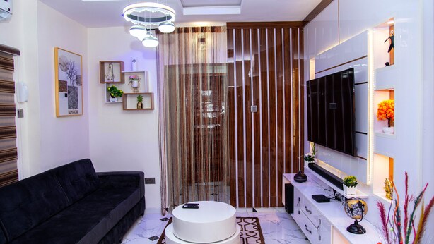 A modern living room with a black sofa, a white coffee table, and a TV mounted on the wall. - Tastefully furnished 2 bedroom apartment with 24/7 Power|WiFI|Security (Alimosho)