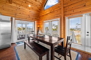 A wooden cabin interior with a dining table, chairs, and a view of the outdoors through large windows.