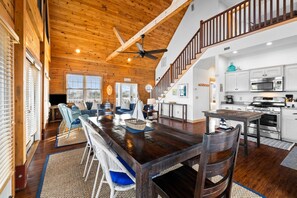 A spacious living area with a wooden ceiling, a dining table with chairs, a kitchen with modern appliances, and a staircase leading to an upper level.