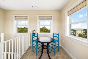 A balcony with a table and chairs, two windows with blinds, a white railing, and a view of buildings and trees.
