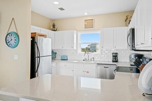Uma cozinha moderna com armários brancos, geladeira de aço inoxidável, lava-louças embutida e uma janela com vista para as casas.