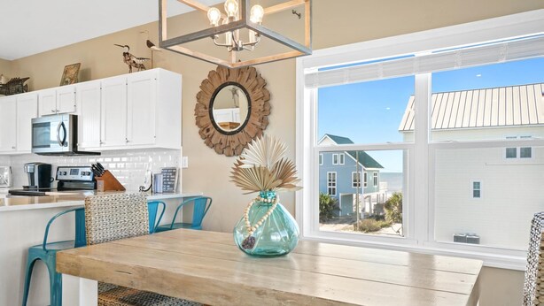 A kitchen with white cabinets, a wooden dining table, and a view of a blue house through a large window.