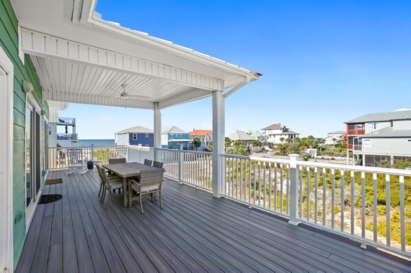 Um deck coberto com mesa e cadeiras, com vista para uma praia e casas ao fundo.