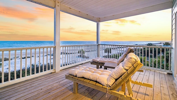 Una terraza de madera con una silla reclinable acolchada y una mesita pequeña, con vista a una playa y una barandilla de madera.