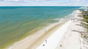 Een strand met helder turquoise water, een zandstrand en enkele mensen die erover lopen. Aan de rechterkant van de afbeelding staan gebouwen, mogelijk onderdeel van een resort of hotel.