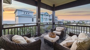 A covered balcony with wicker furniture and a view of coastal houses during sunset.