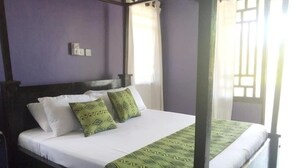 Una cama con dosel, ropa de cama blanca y almohadas estampadas en verde, frente a una pared morada con una ventana y cortinas.