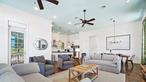 Un salón amplio con un sofá gris grande, un sillón gris más pequeño y una mesa de centro de madera. En el fondo se ve una zona de comedor con una mesa y sillas.