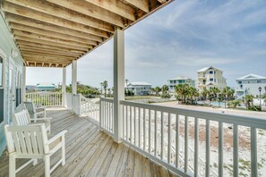 A wooden deck with white railings and chairs, overlooking a beach and buildings.