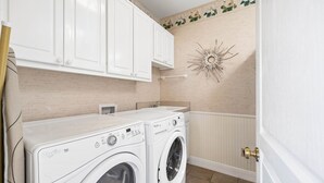 A laundry room with two front-loading Whirlpool Duet washers, white cabinetry, and a decorative sunburst wall art piece.