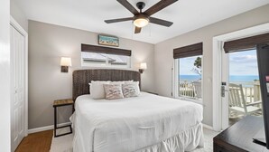 A bedroom with a bed, a ceiling fan, a nightstand, a lamp, a window with a view of the beach, and a door.