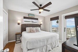 A bedroom with a bed, a ceiling fan, a nightstand, a lamp, a window with a view of the beach, and a door.