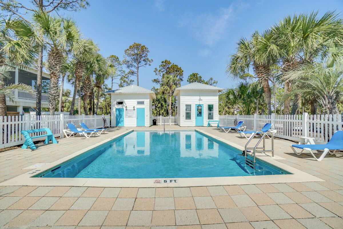 A rectangular swimming pool with a 5 ft depth marker, surrounded by a white fence, blue lounge chairs, and palm trees.