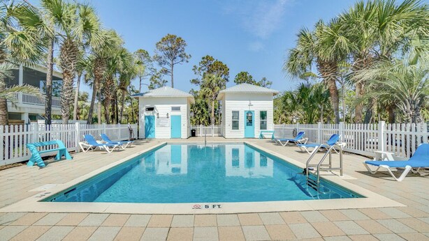 A rectangular swimming pool with a 5 ft depth marker, surrounded by a white fence, blue lounge chairs, and palm trees.