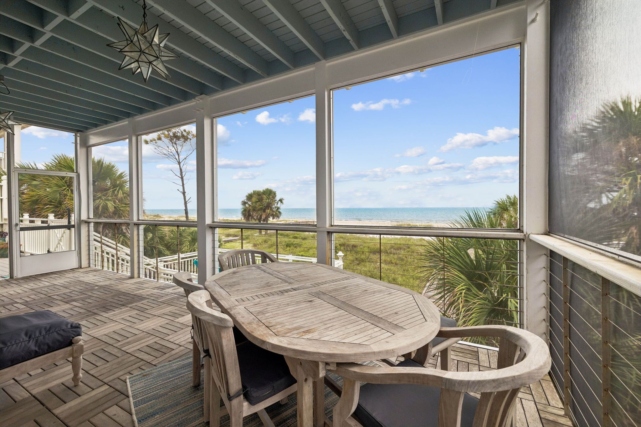 A covered outdoor area with a wooden table and chairs, large windows offering a view of the beach and ocean.