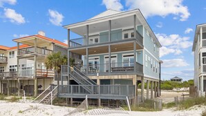 A multi-story beachfront house with a balcony, a staircase, and a sandy beach in the background.