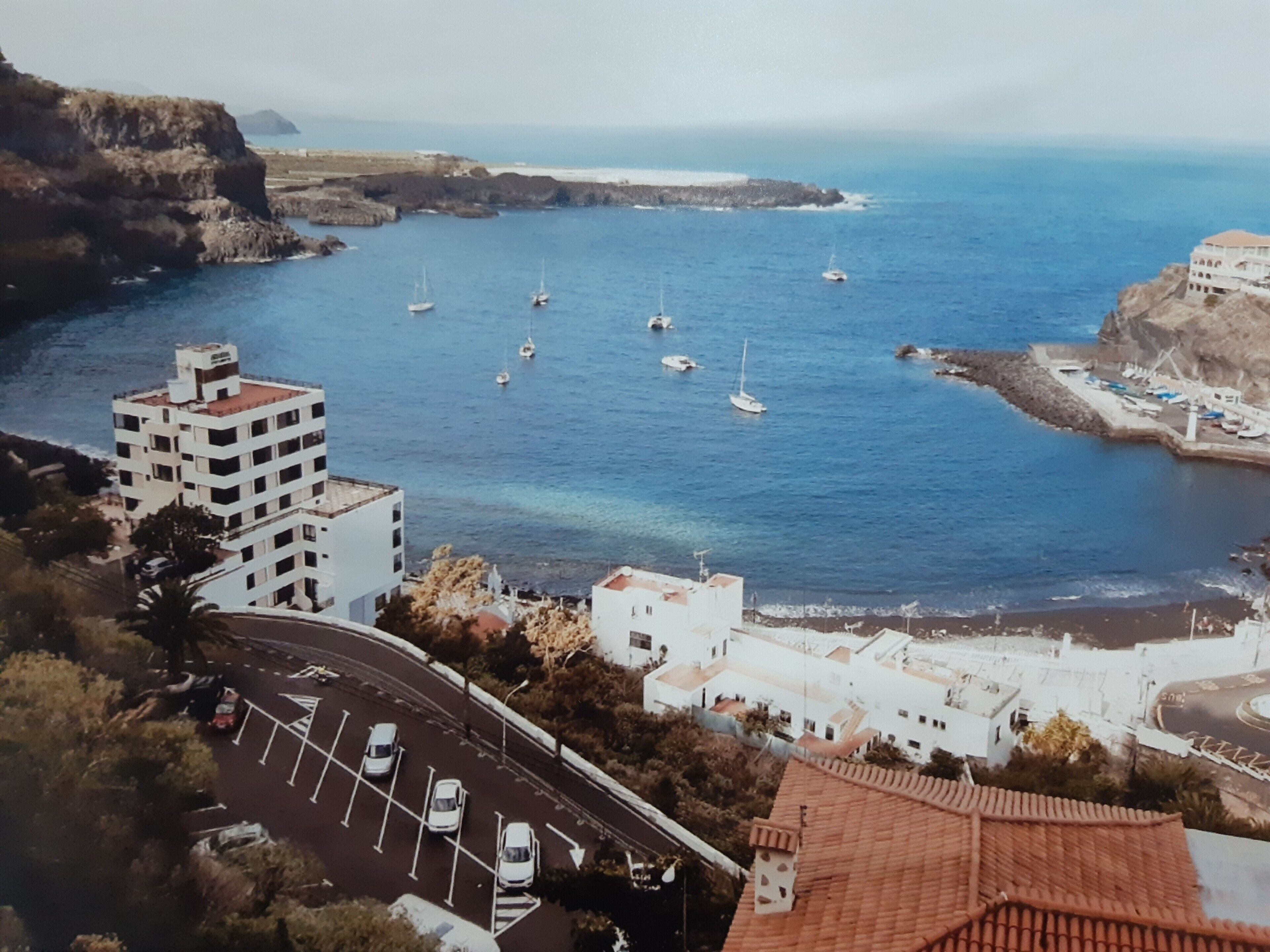 A coastal town with buildings, a road with parked cars, and boats in the water.