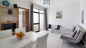 Ein modernes Wohnzimmer mit einer Couch, Sesseln, einem Esstisch mit Obst, einem Fernseher und einem Bücherregal.