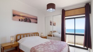Ein Schlafzimmer mit Bett, Nachttischen, einem Fenster mit Vorhängen, Meerblick und einem Sonnenuntergangsbild an der Wand.