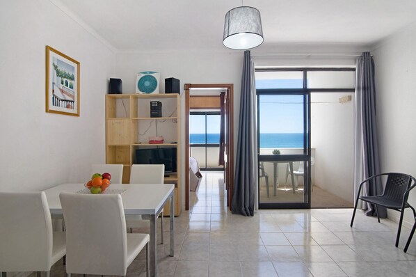A room with a dining table, chairs, a bookshelf, a ceiling fan, and a view of the sea through a large window.