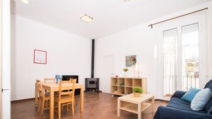 Una sala de estar con un sofá, una mesa de comedor con sillas, una pequeña mesa de madera con una planta, una estantería, una chimenea y un amplio ventanal con vistas.