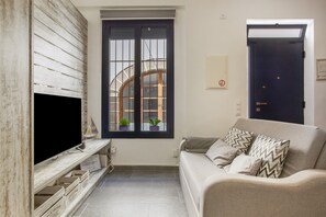 A living room with a flat-screen TV, a sofa with patterned cushions, a window with a view of a building, and a door. - Spectacular designer loft just five minutes' walk from the beach (Canet de Mar)