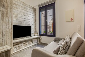 A living room with a TV on a wooden stand, a sofa, and a window with plants.