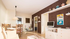 A living room with a sofa, a coffee table, a TV, a dining area with chairs, a white cabinet, and a wall with decorative items. - Apartment with Sea View, Terrace, and Beachfront Location (Canet  de mar)
