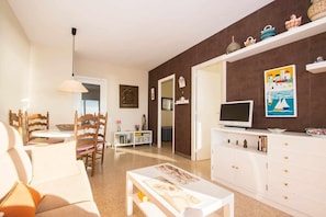 A living room with a sofa, a coffee table, a TV, a dining area with chairs, a white cabinet, and a wall with decorative items.